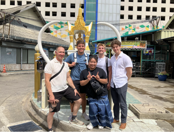 One professor and four students posing for a photo at a market in Thailand. One person is making a "W" with their hands in the center.